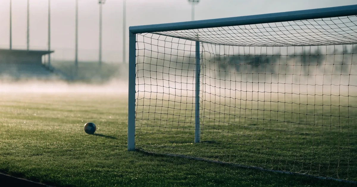 Ein Fußballtor mit gespanntem Netz steht auf einem leeren Rasenplatz, davor ein einzelner Ball, Morgendunst liegt über dem Feld.