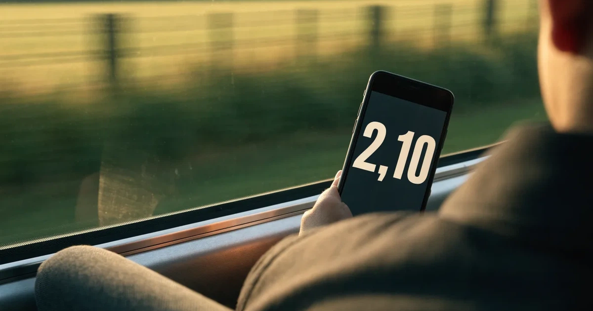 Eine Hand hält ein Smartphone mit dunklem Display in einem Zug, am Fenster ziehen verschwommen Felder vorbei.