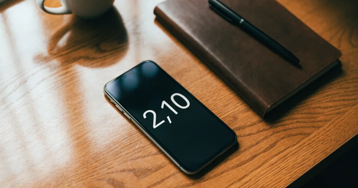Ein Holzschreibtisch im Morgenlicht mit geschlossenem Notizbuch, Kugelschreiber und Kaffeetasse, daneben ein Smartphone mit dunklem Bildschirm.