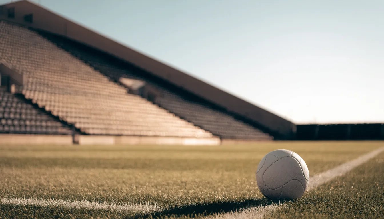Ein Fußball liegt am Spielfeldrand eines Rasenplatzes, im Hintergrund eine leere Tribüne unter hellem Himmel.