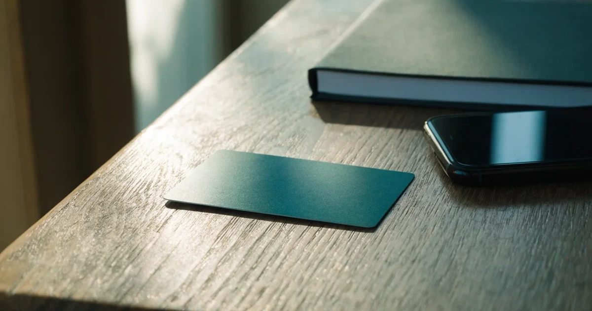 Eine Plastikkarte ohne Aufdruck liegt auf einem Holztisch neben einem Smartphone mit dunklem Bildschirm und einem Notizbuch.