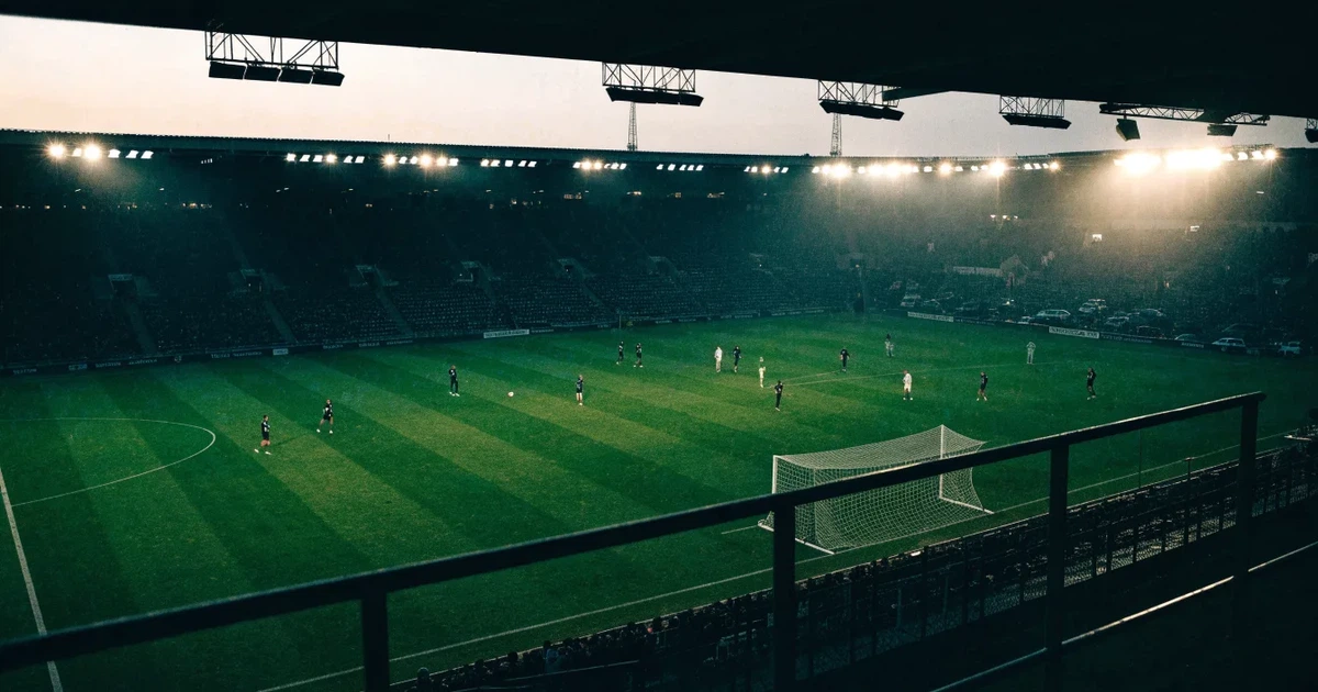 Ein Fußballstadion unter Flutlicht aus großer Entfernung, davor ein dunkler Parkplatz und ein heller Abendhimmel.
