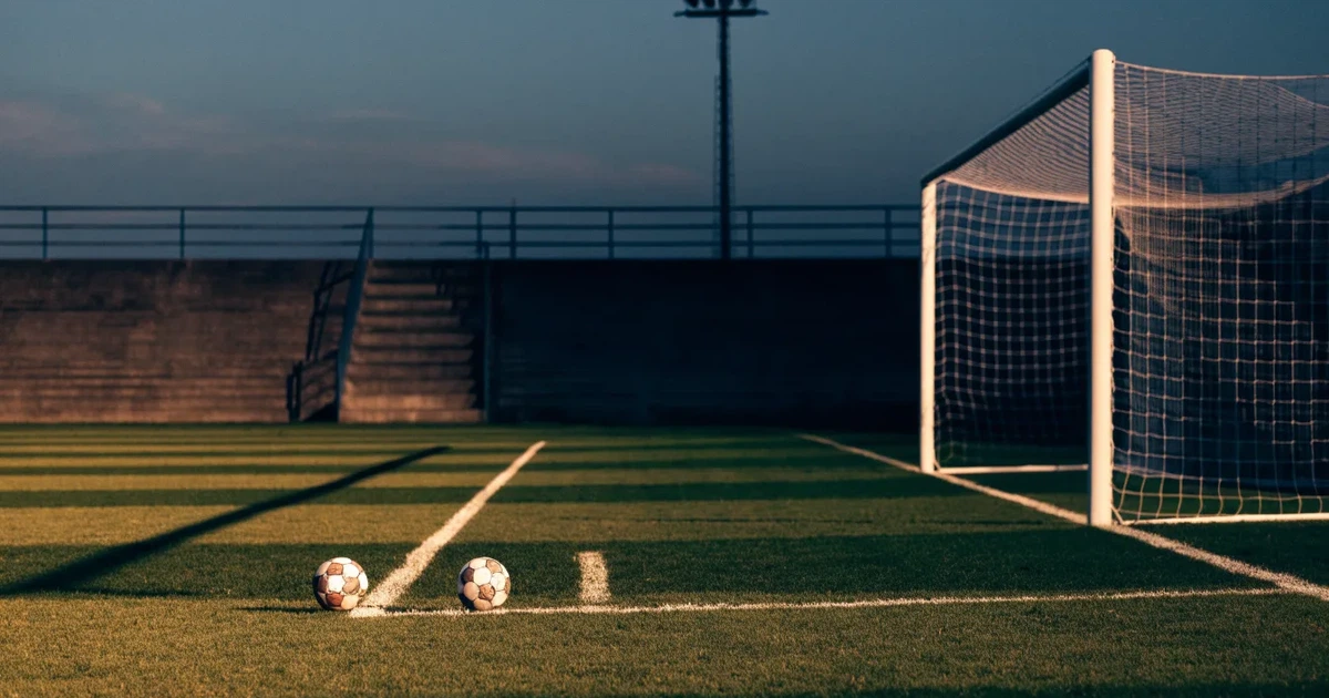 Ein Fußballplatz mit frisch gemähtem Rasen im Abendlicht, ein Ball liegt im Strafraum, im Hintergrund unscharf eine leere Tribüne.
