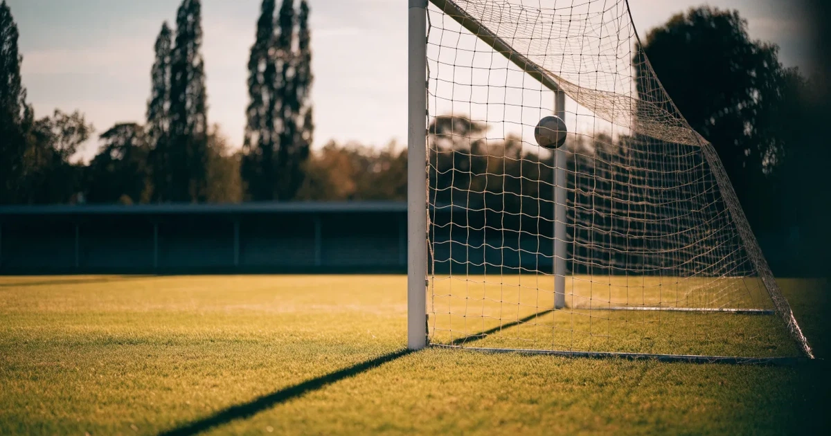 Ein Fußballtor mit Netz auf einem leeren Platz im Abendlicht, der Rasen frisch gemäht, im Hintergrund Zypressen.