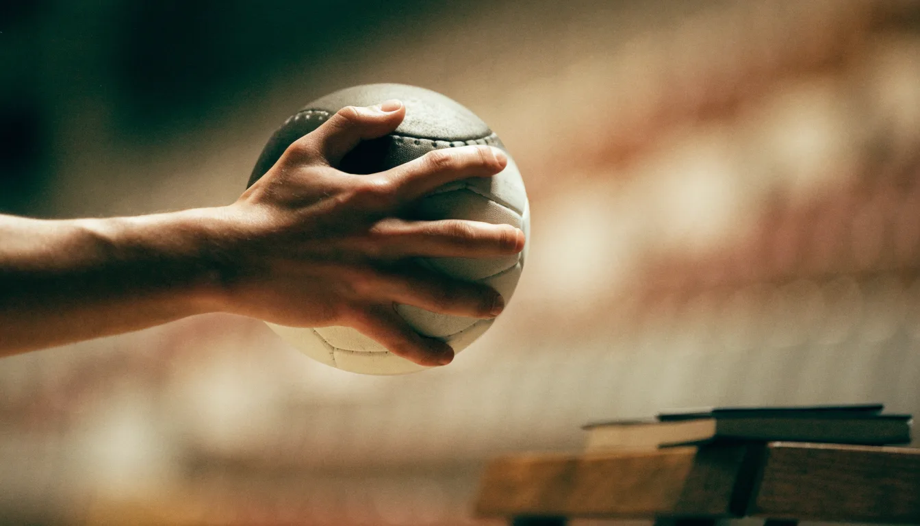 Eine Hand hält einen Handball, im Hintergrund eine unscharfe Tribüne, daneben liegt ein Notizblock auf einer Bank.