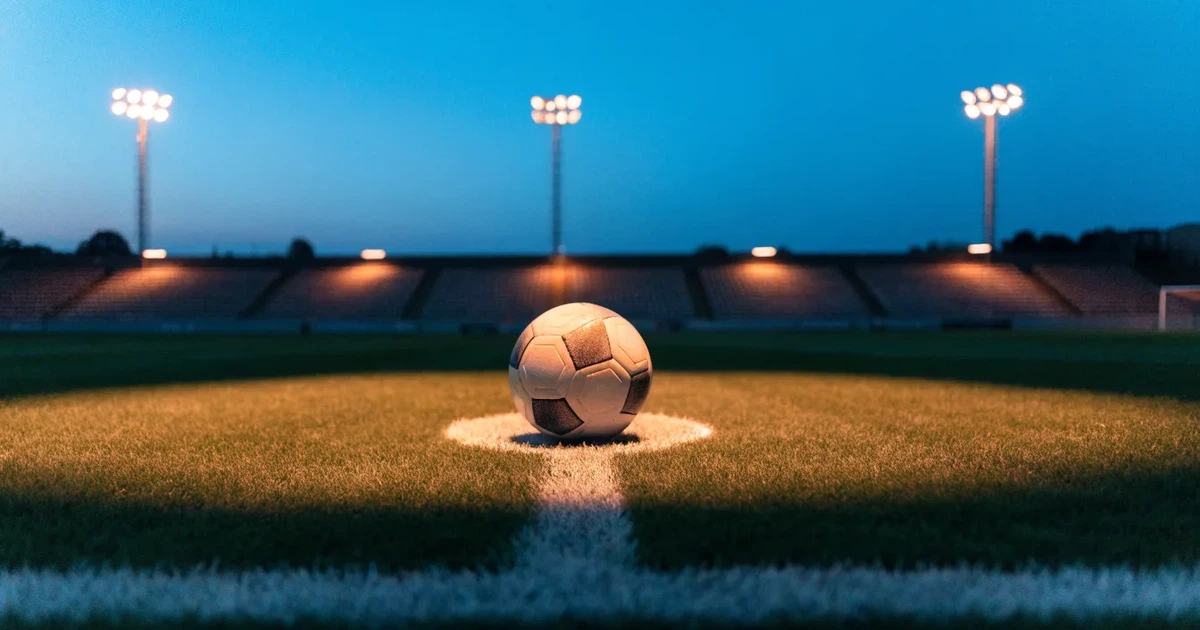 Ein Fußball liegt auf dem Anstoßpunkt eines leeren Rasenplatzes, im Hintergrund eine Tribüne und Flutlichtmasten in der Abenddämmerung.