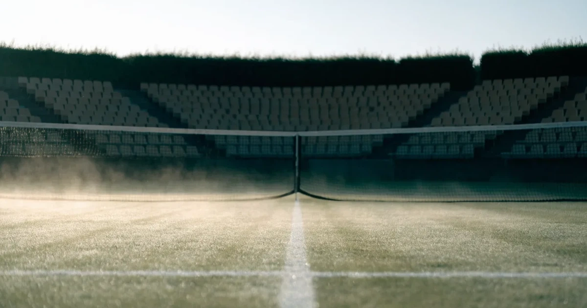 Ein Tennisplatz mit Rasenbelag im Morgenlicht, Netz und weiße Linien, im Hintergrund eine ruhige Tribüne.