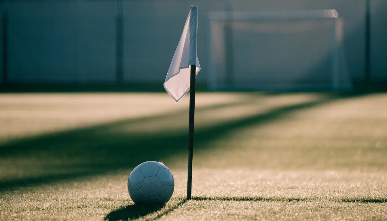 Ein Fußball liegt auf Rasen neben einer Eckfahne ohne Aufdruck auf einem leeren Platz im Morgenlicht.