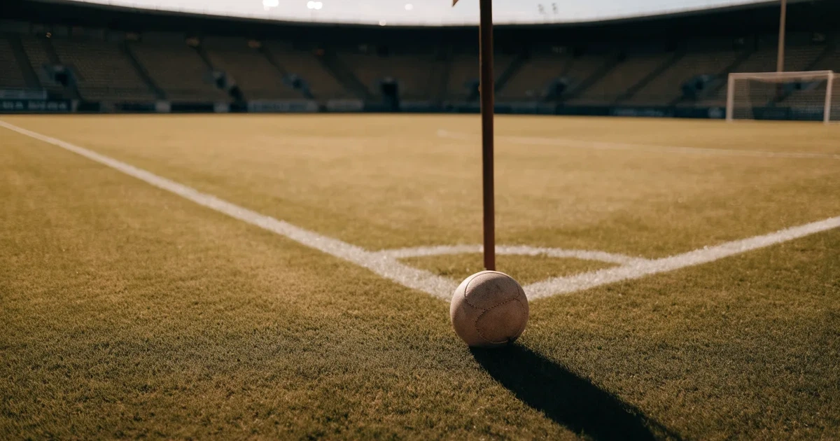 Ein Fußball liegt am Viertelkreis an der Eckfahne eines Rasenplatzes, im Hintergrund leere Tribünen in warmem Abendlicht.