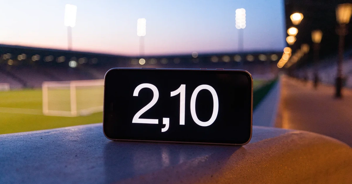 Ein großes Fußballstadion mit hell erleuchtetem Rund, aus der Ferne am Abend gesehen, davor eine breite Allee mit Laternen.