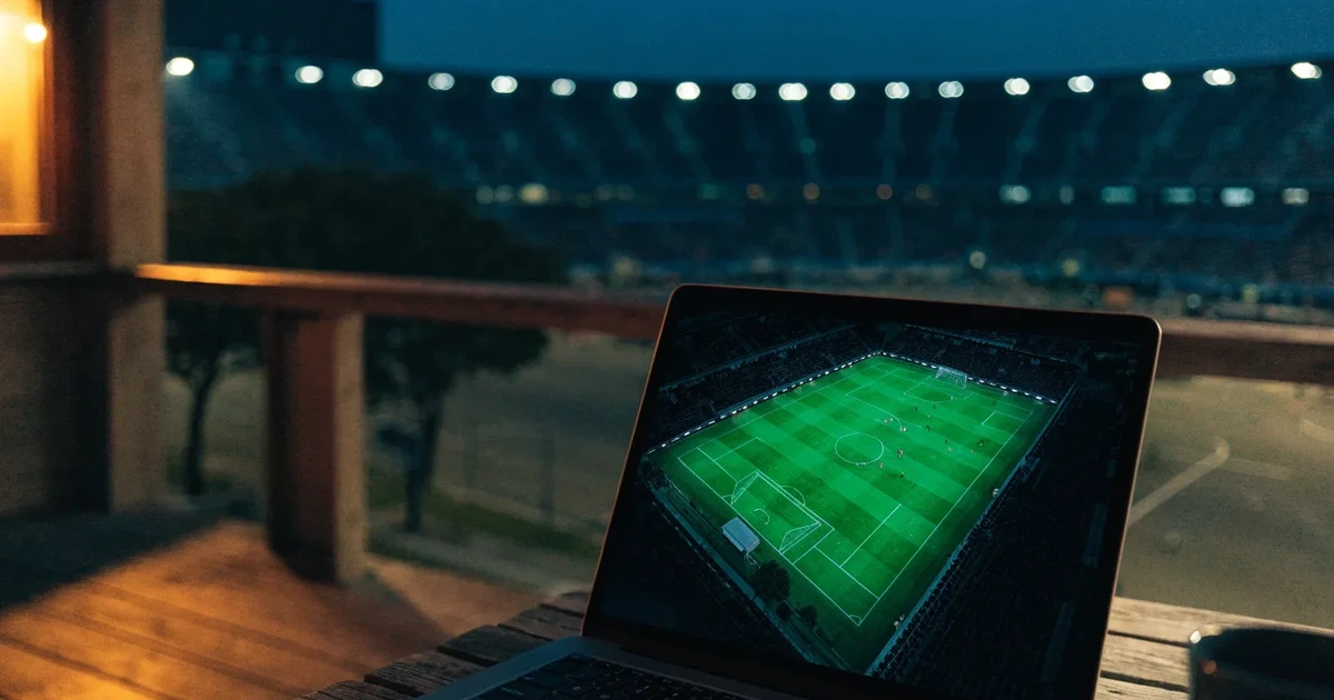 Ein hell erleuchtetes Stadion aus der Ferne bei Nacht, davor eine stille Straße und ein paar Bäume.